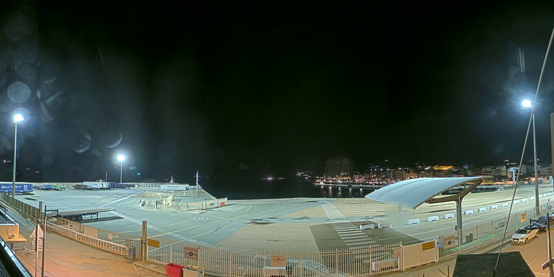 An empty, brightly lit port at night overlooks a dark body of water with a distant, faintly lit town.