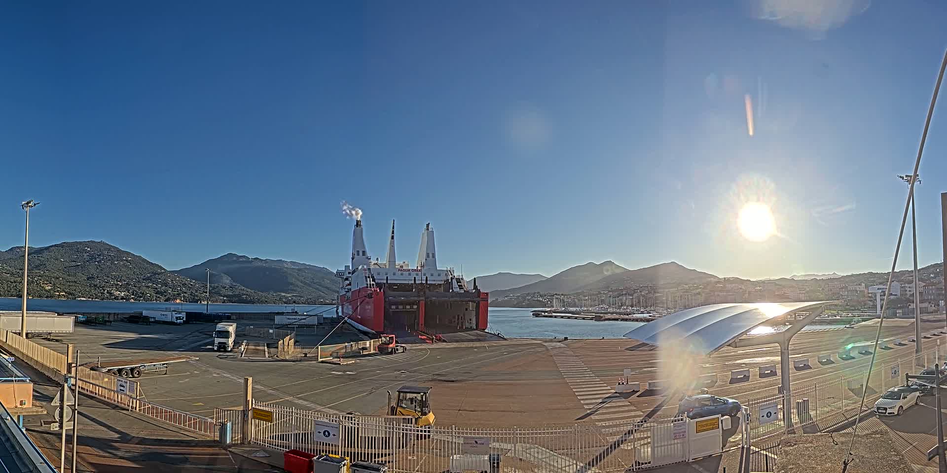 A large red and white cargo ship is docked at a port, with mountains and a bright sun in the background on a clear day.