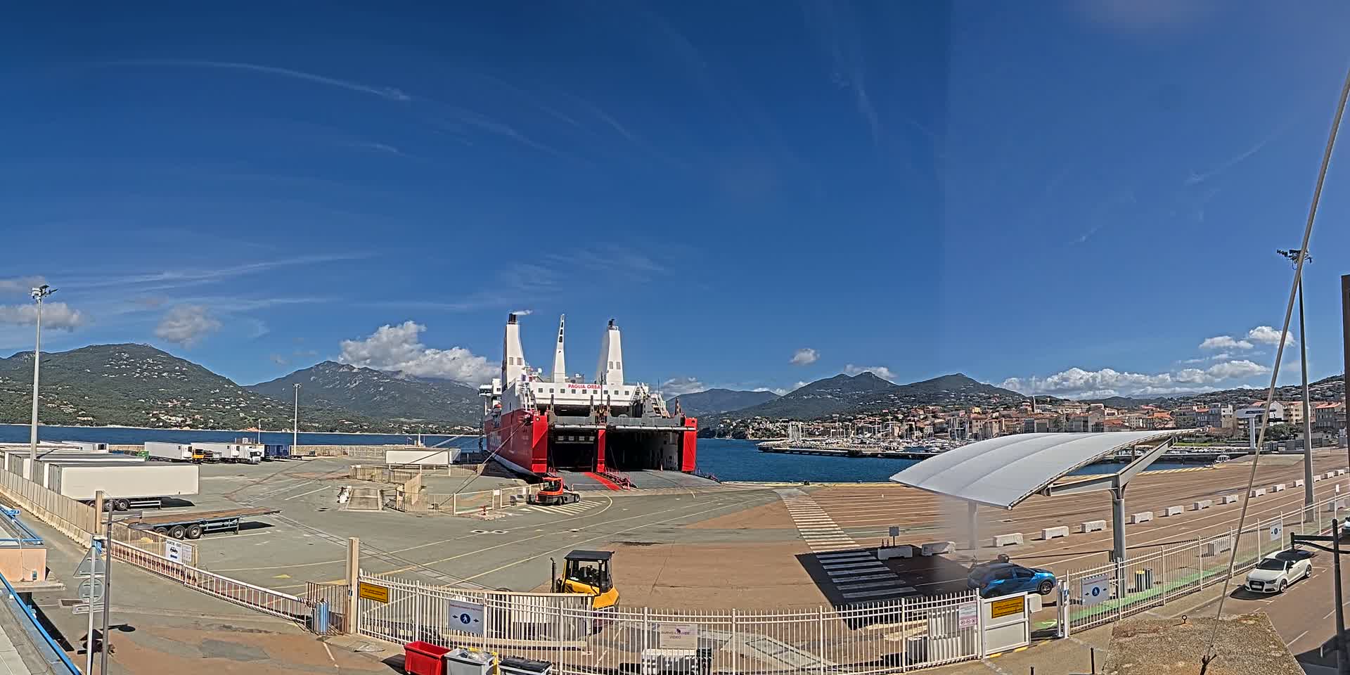 A large red and white ferry is docked at a port under a clear blue sky with a few scattered clouds, mountains in the background.