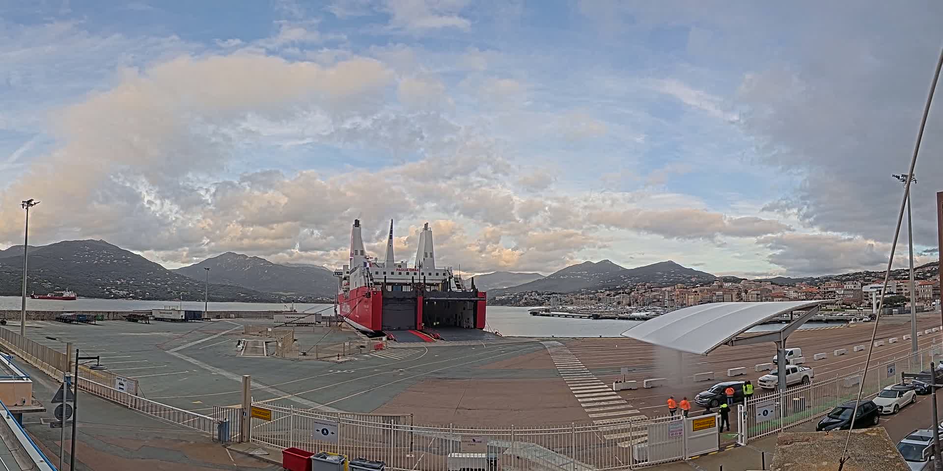 A large red and white ferry is docked at a port under a partly cloudy sky, with mountains and a town visible in the background.