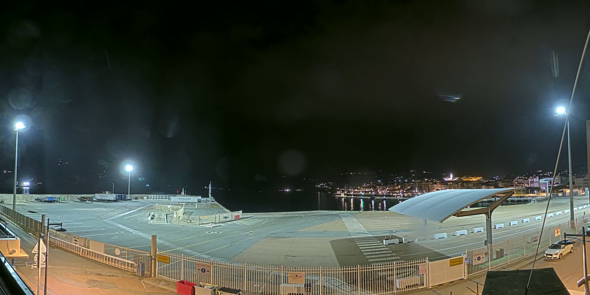 A nighttime view of an empty port area with a few vehicles, illuminated by artificial lights, overlooking a calm body of water and a distant town.