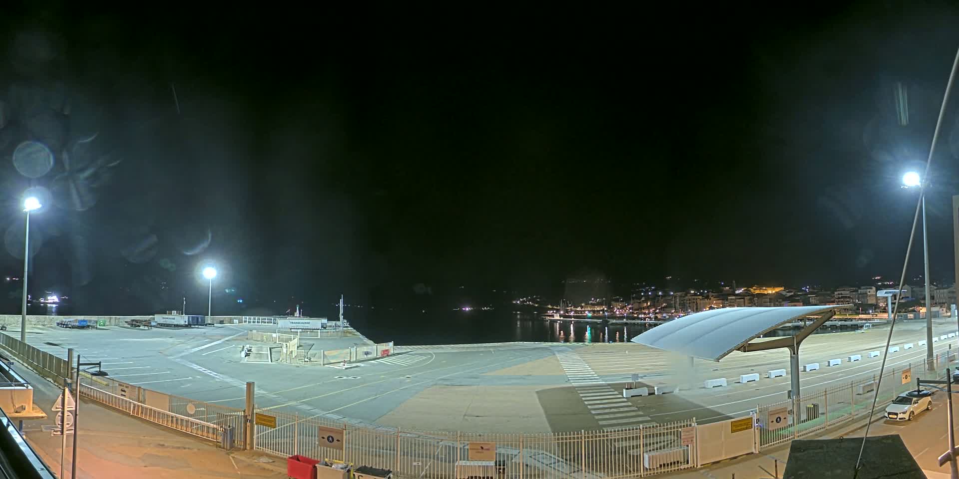 A nighttime view of an empty port area with a covered waiting area, illuminated by several bright lights, overlooking a dark body of water and a dimly lit town in the distance.