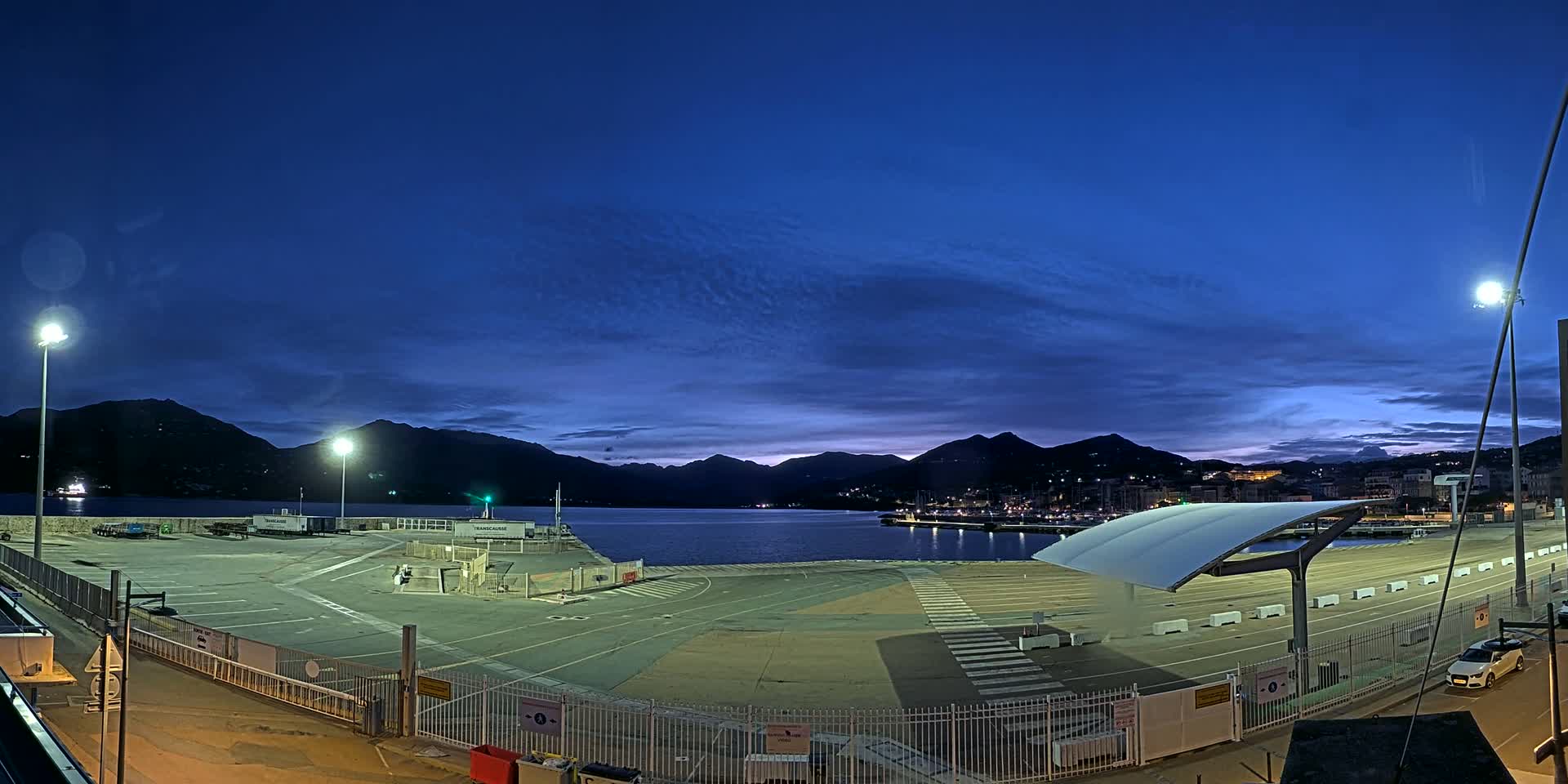 An empty, illuminated port area at twilight, with mountains and a calm body of water in the background under a clear, dark blue sky.