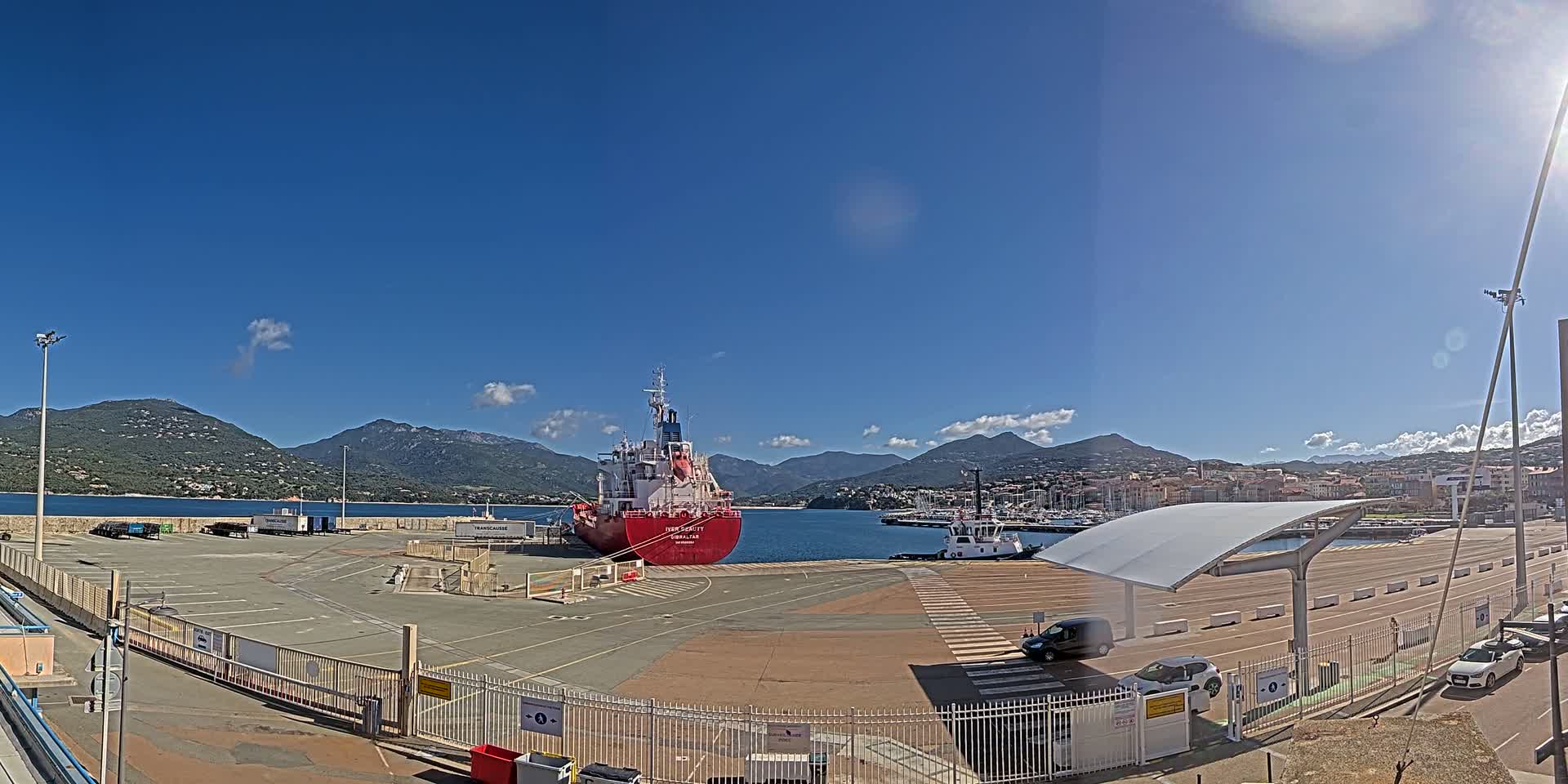 A red cargo ship is docked at a port on a sunny day, with mountains and a town visible in the background.