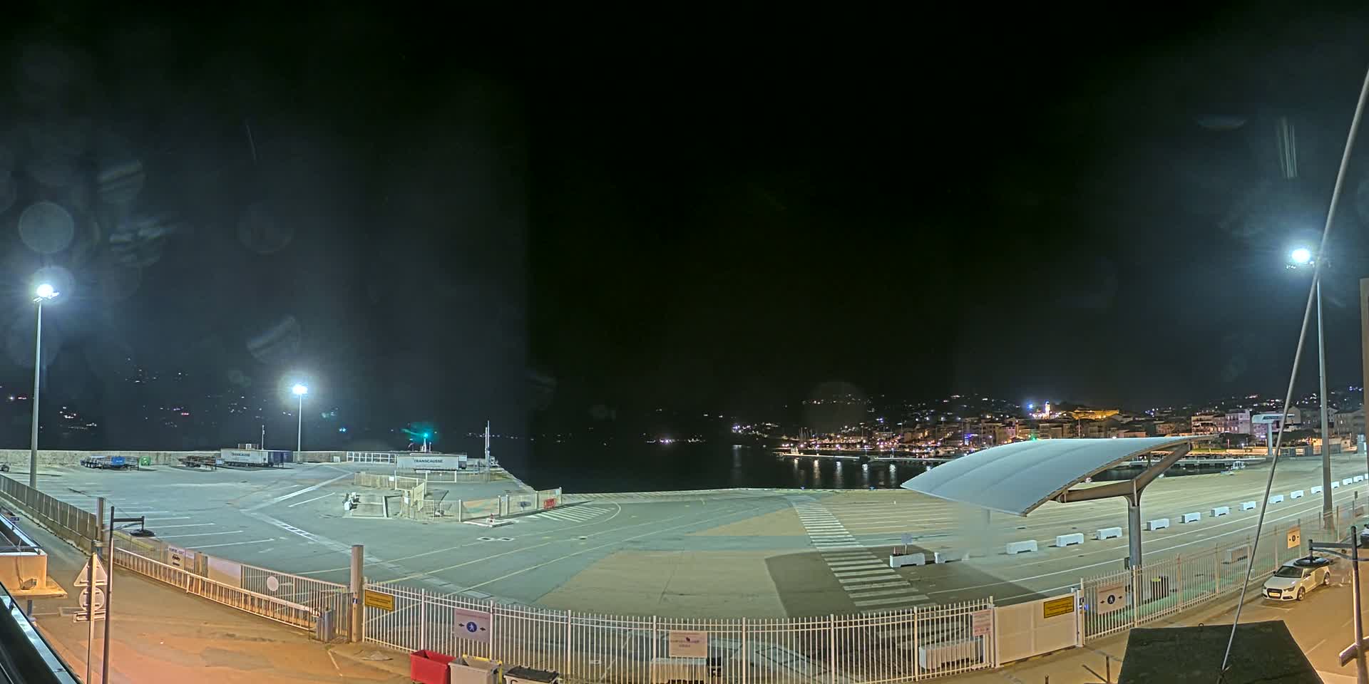 A nighttime view of an empty port area with a few parked vehicles, illuminated by artificial lights, overlooking a city skyline across a dark body of water.