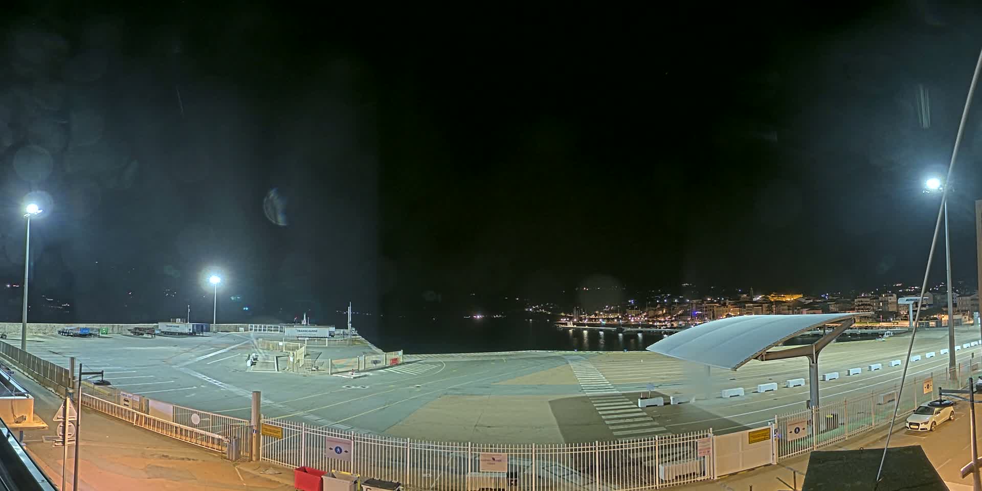 An empty, brightly lit port area at night overlooks a dark body of water with a town faintly visible in the distance.