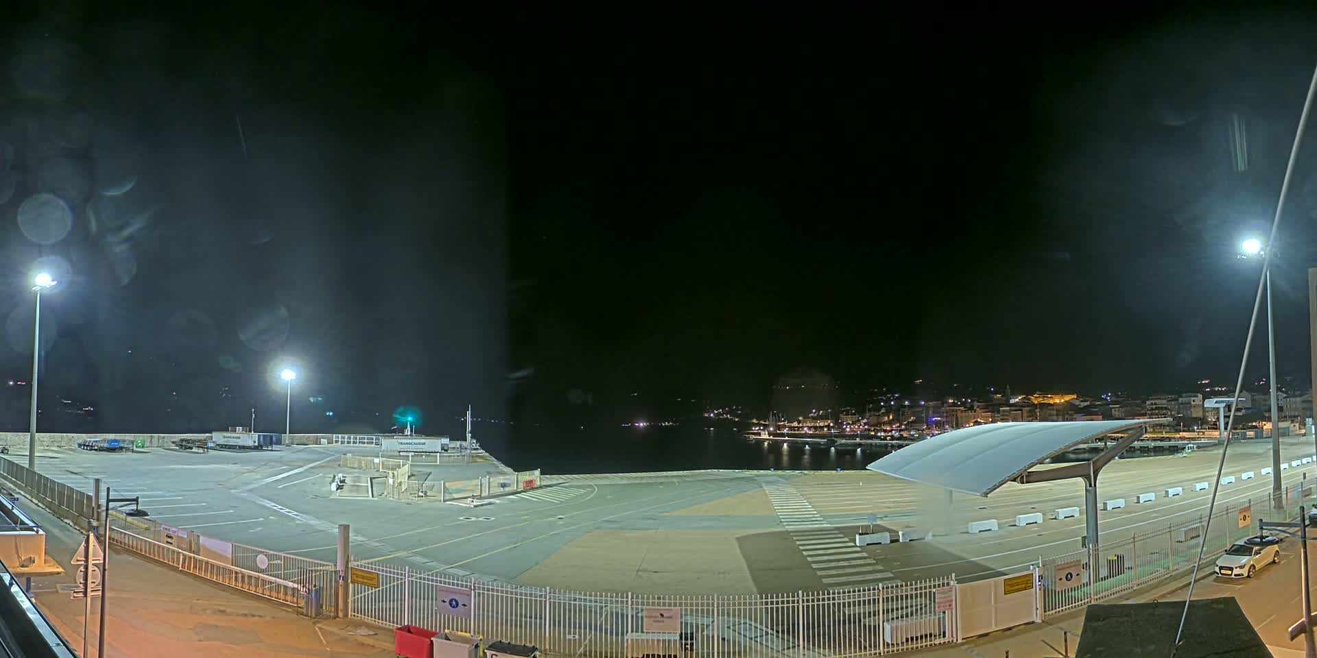 A nighttime view of an empty port area with a few cars parked, illuminated by several bright lights, overlooking a calm body of water with a town in the distance.