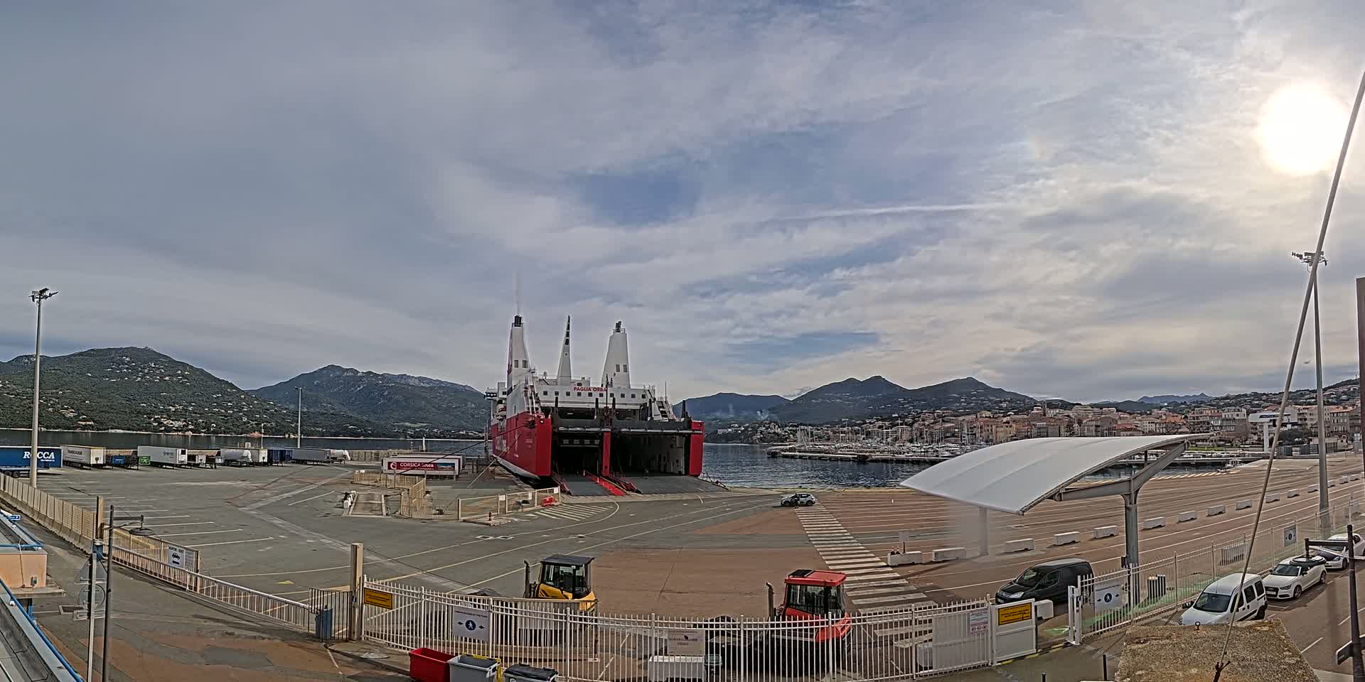 A large red and white ferry is docked at a port, with mountains in the background under a partly cloudy sky.