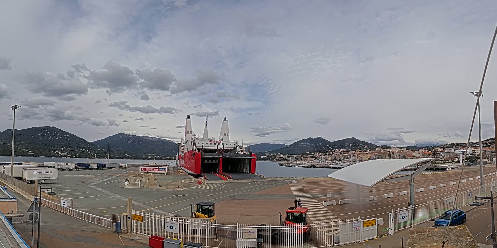 A large red and white ferry is docked at a port, with overcast skies and mountains in the background.