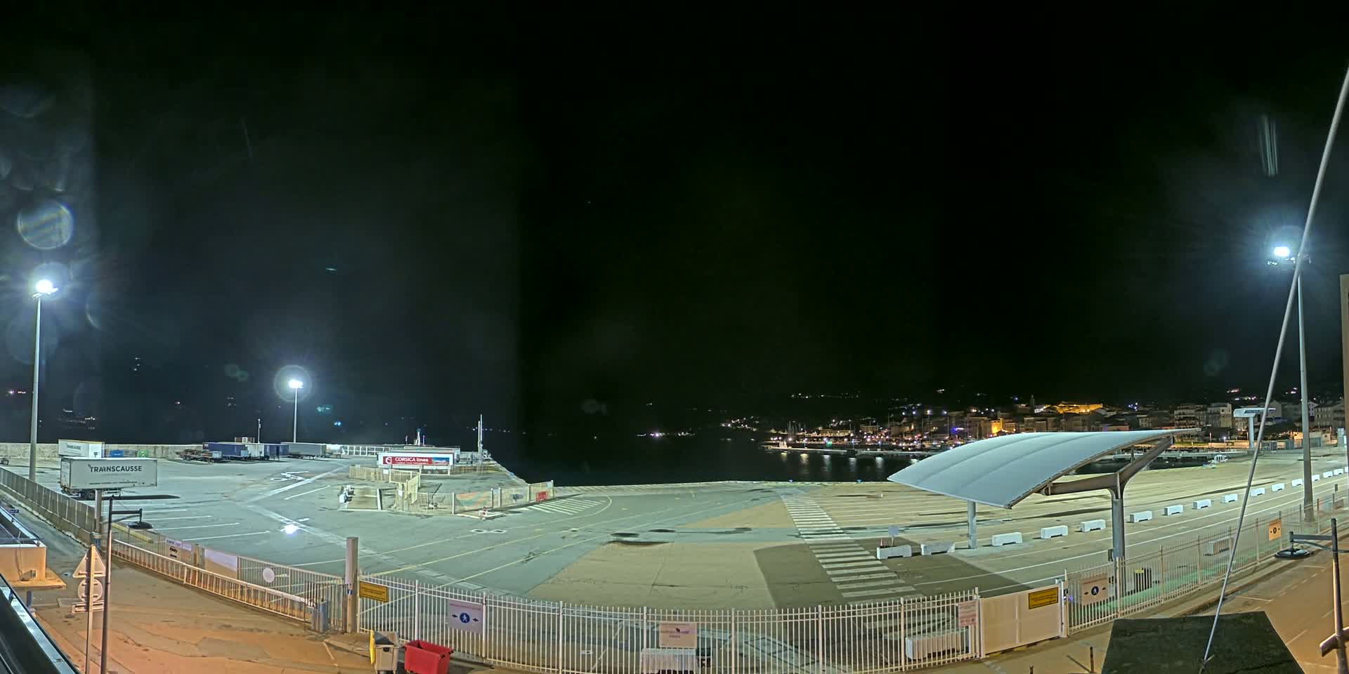 A nighttime view of a port area with several trailers, a large empty paved lot, and a city skyline visible in the distance under a clear, dark sky.