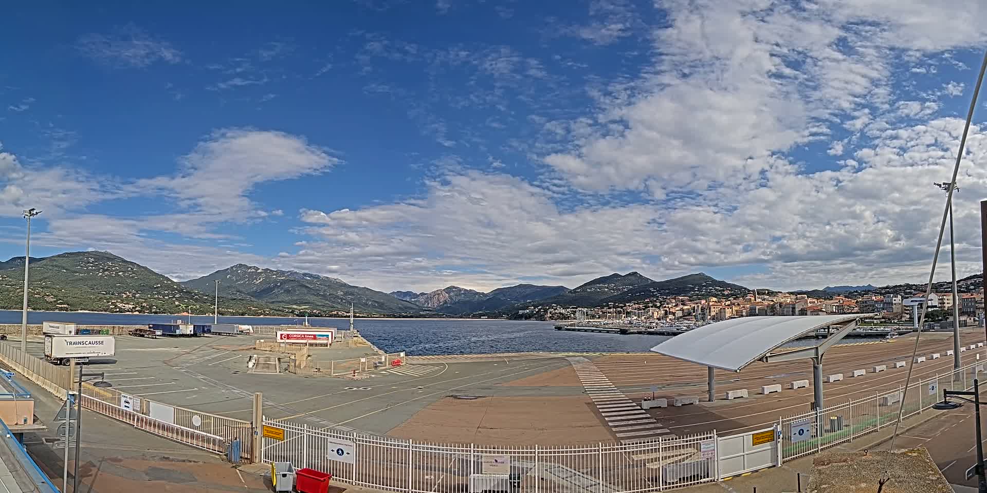 A mostly sunny day reveals a port area with mountains in the background, a body of water in between, and several docked boats.