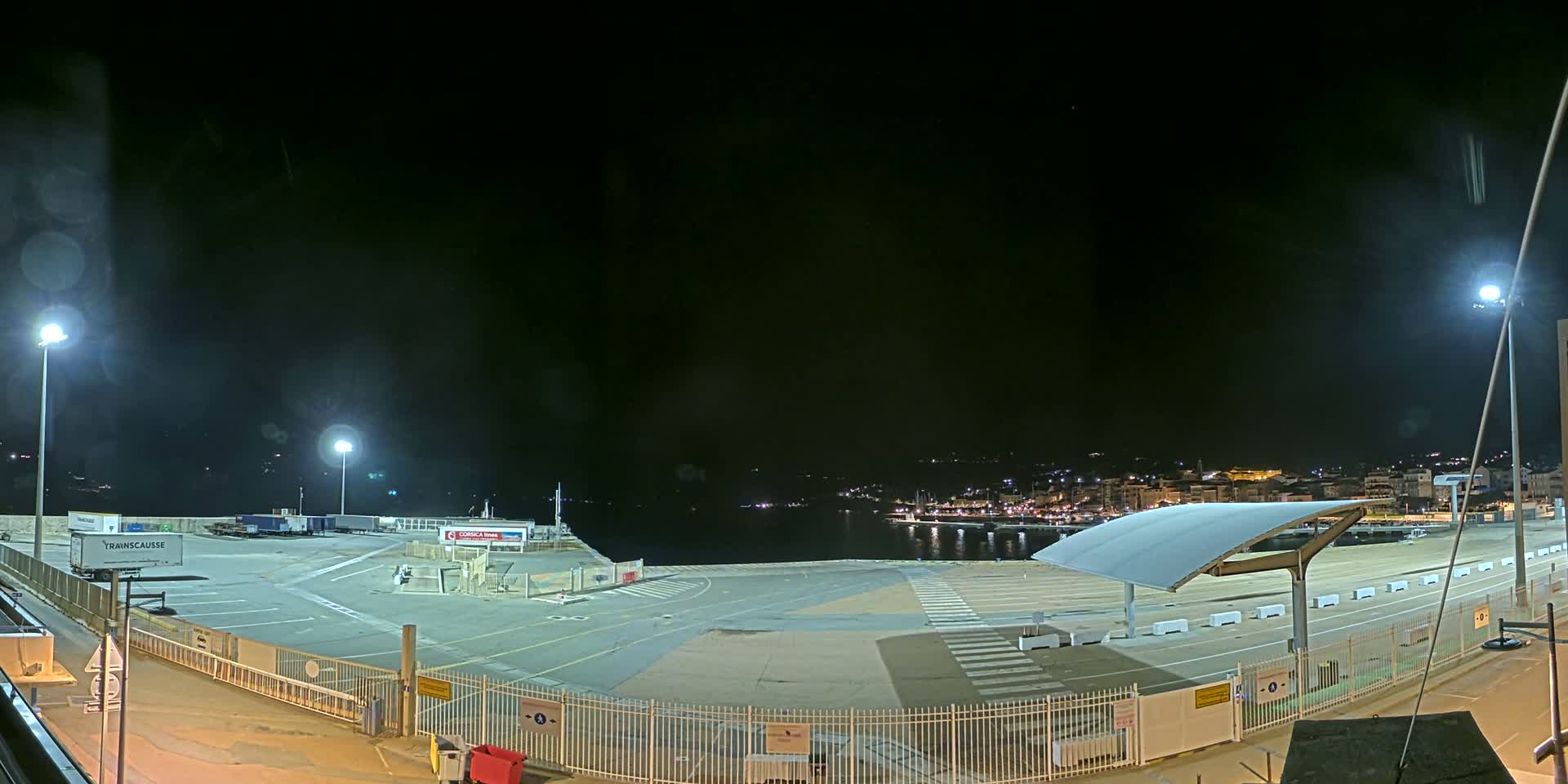 An empty, brightly lit port area at night overlooks a dark, calm body of water with a town visible in the distance.