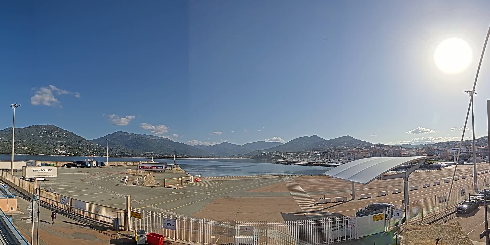 A sunny day at a port, showing a large empty paved area enclosed by fences, with mountains and a town visible in the distance across a body of water.