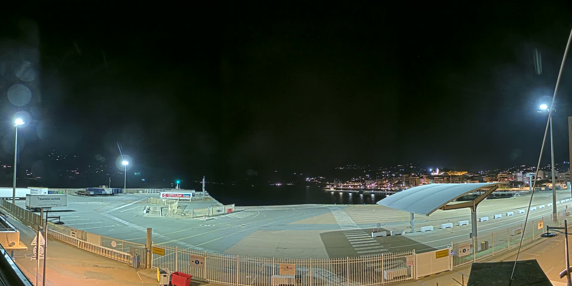 An empty, brightly lit port area at night, with a town visible across the water.