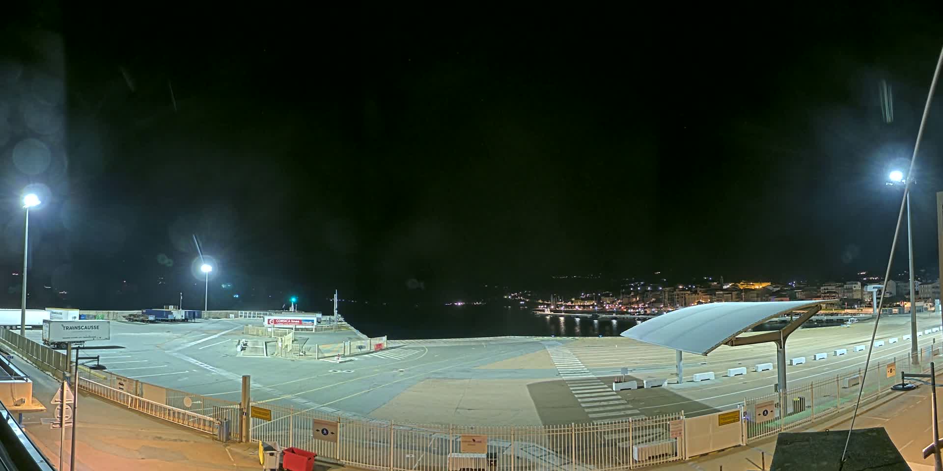 A nighttime view of an empty port area with a few parked trucks and a distant, illuminated town across the water under a clear, dark sky.