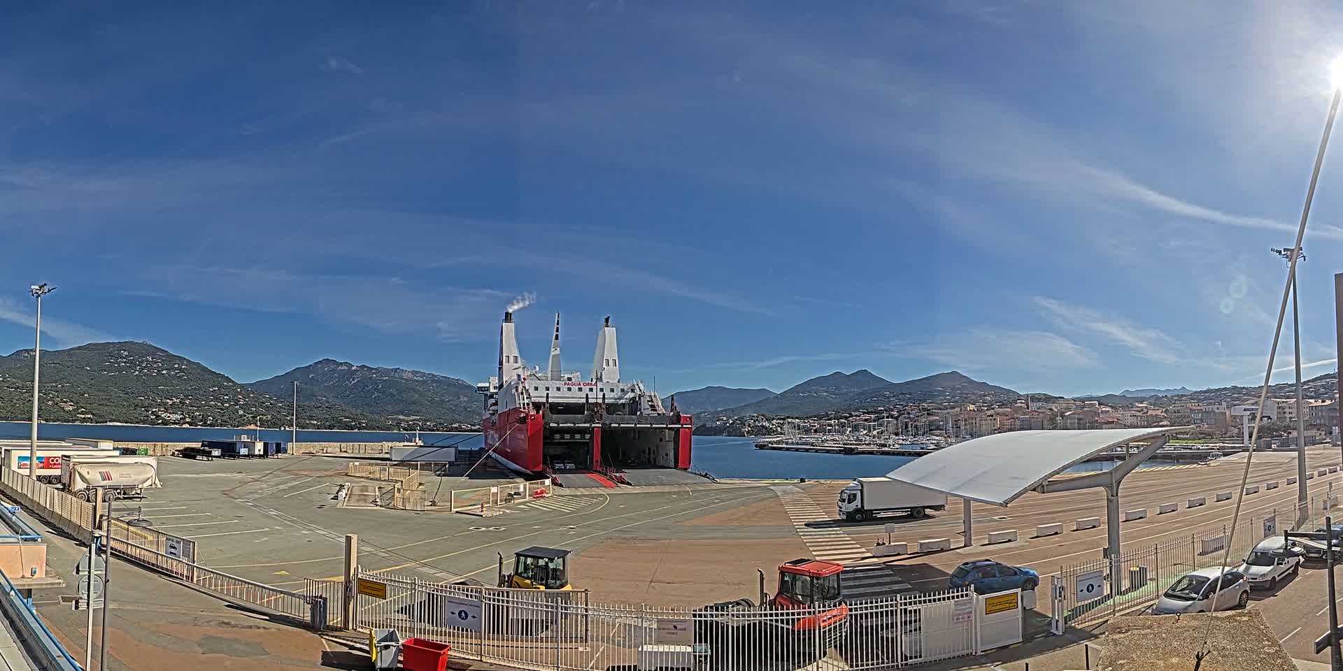 A large red and white ferry is docked at a port under a clear blue sky, with mountains in the background and several vehicles parked nearby.