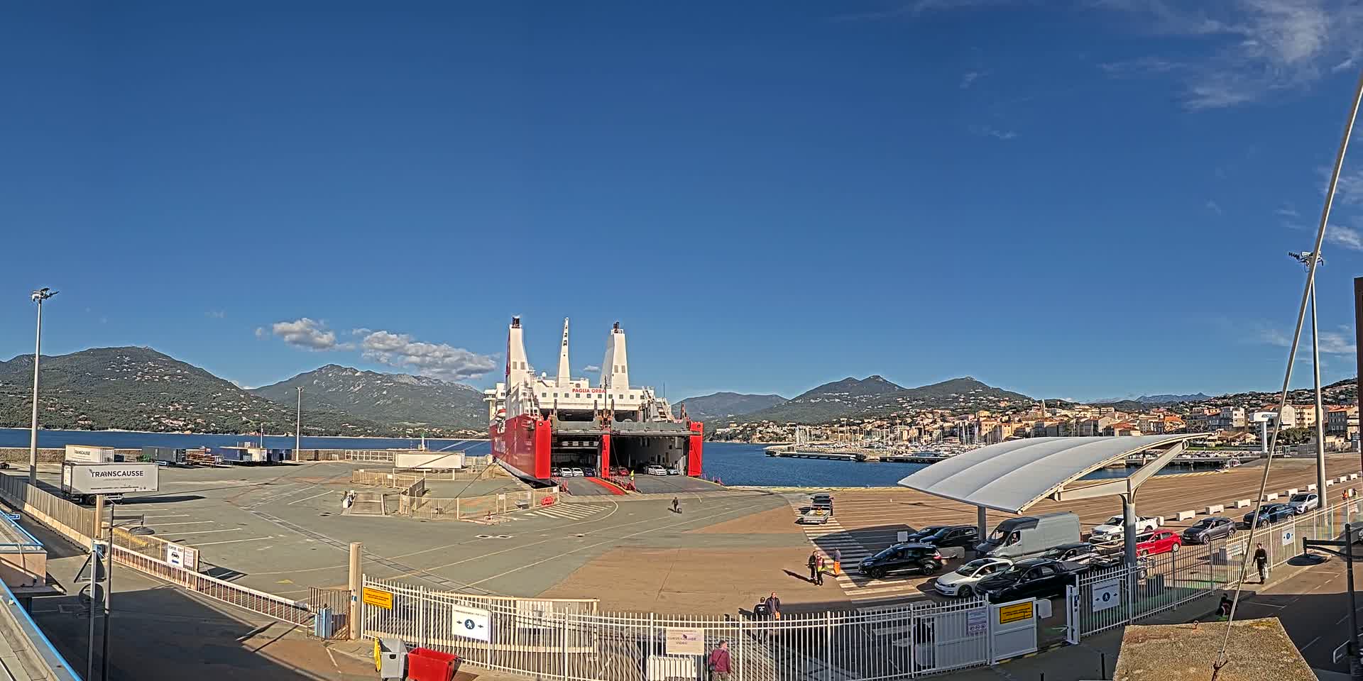 A large red and white ferry is docked at a port, under a clear blue sky with a few wispy clouds, and cars are being loaded and unloaded.