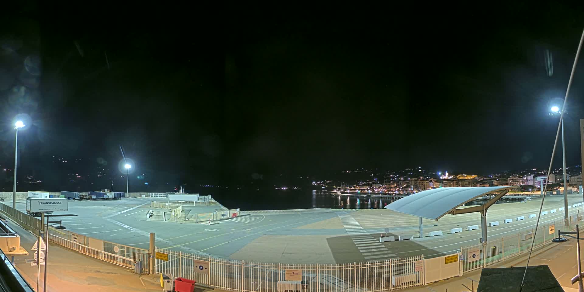 An empty, brightly lit port area at night, with a town visible across the water in the background under a clear, dark sky.