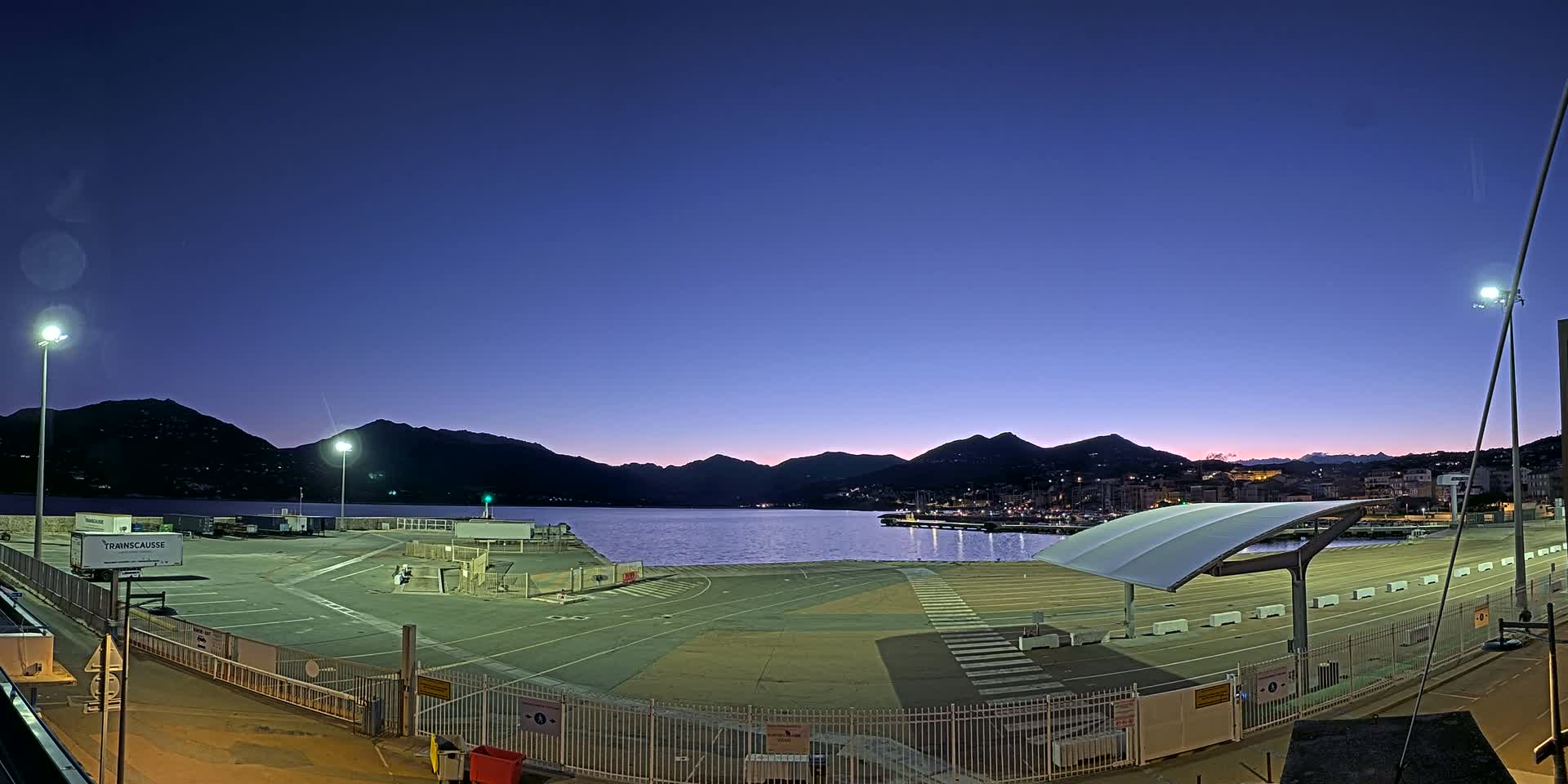 An empty, illuminated port area at twilight, bordered by a fence, with mountains and calm water visible in the background under a clear, dark blue sky.