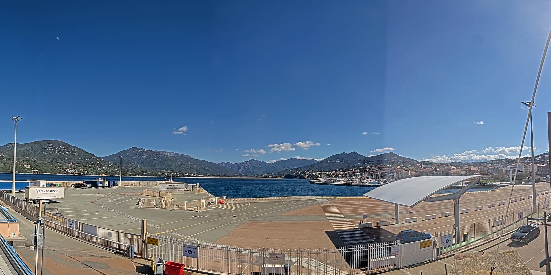 A mostly empty port area with a few cars and trucks, under a clear blue sky, overlooks a calm body of water and mountains in the distance.