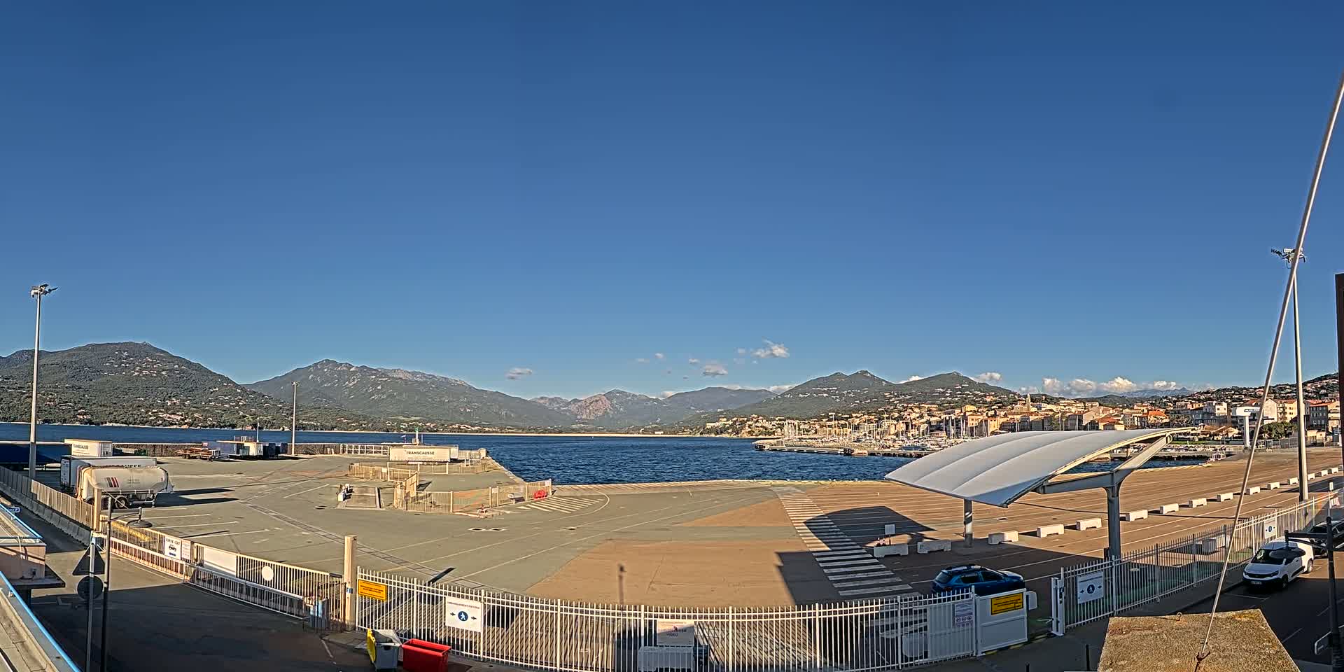 An empty port area with a few cars and trucks is situated next to calm blue water, backed by mountains under a clear blue sky.