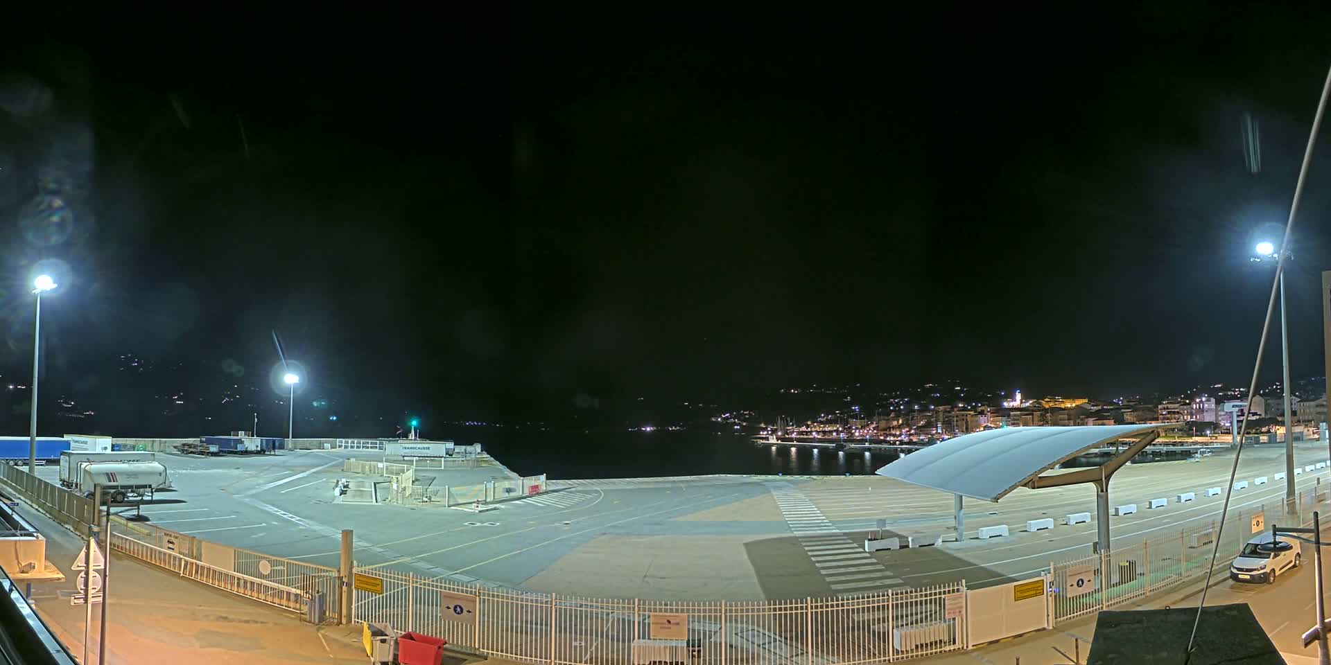 A nighttime view of an empty port area with several parked trucks and a single car, illuminated by artificial lights, overlooking a calm body of water and a distant city skyline.