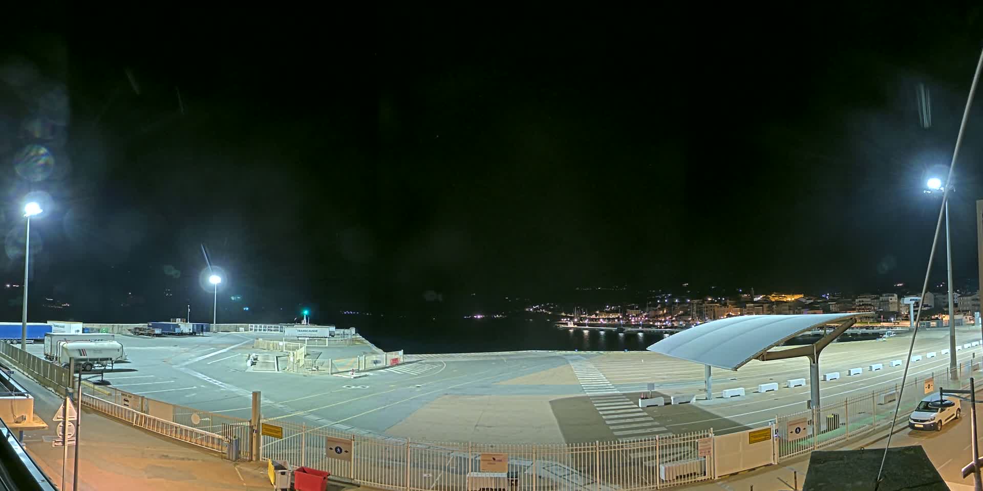 A nighttime view of an empty port area with several parked trucks and a distant town illuminated under a dark, clear sky.