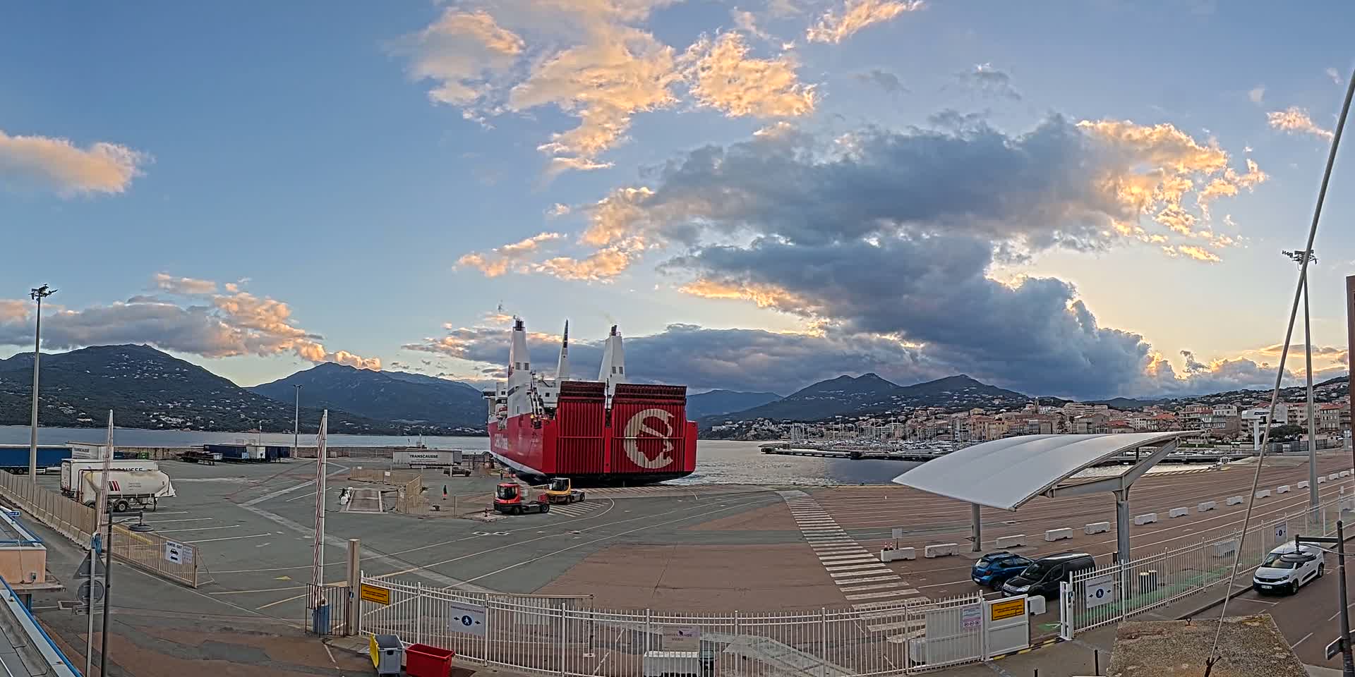 A large red ferry is docked in a port, under a partly cloudy sky with hints of sunset,  with mountains in the background.