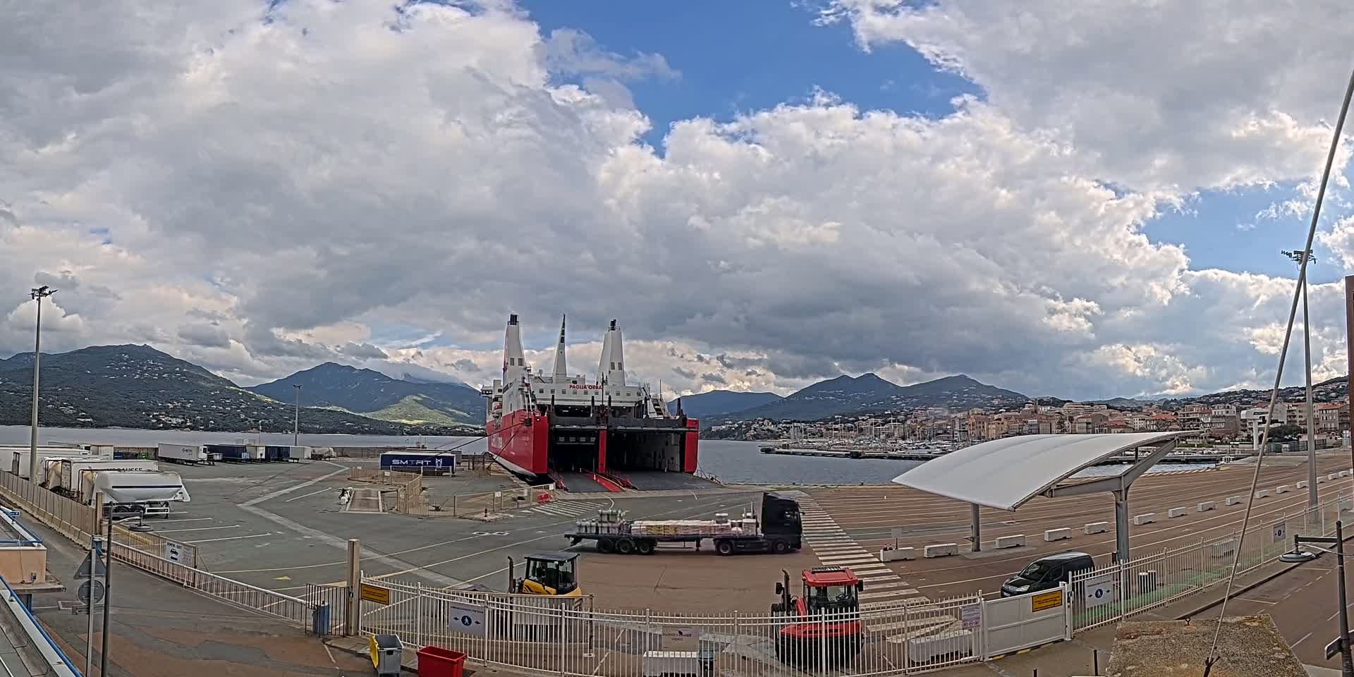 A large red and white ferry is docked at a port under a partly cloudy sky, with mountains and a town visible in the background.