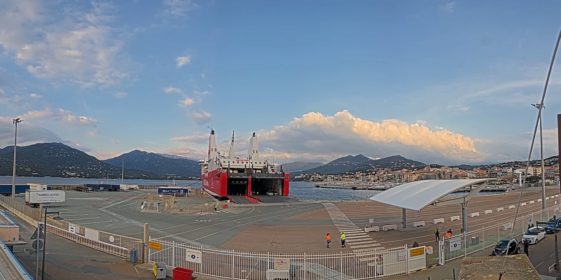 A large red and white ferry is docked at a port, with mountains in the background under a partly cloudy sky.