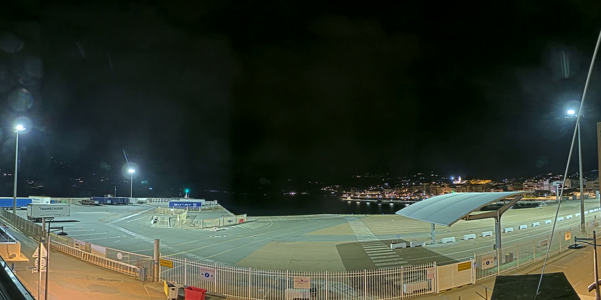 At night, a brightly lit port area shows empty parking spaces, semi-trucks, and a distant city skyline under a dark, clear sky.