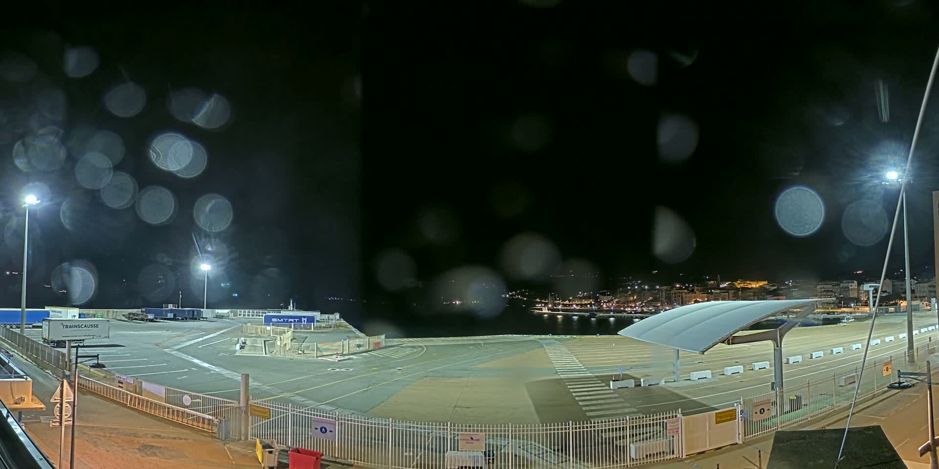 A nighttime view of an empty port area with rain on the lens, showing a harbor and distant lights across the water.