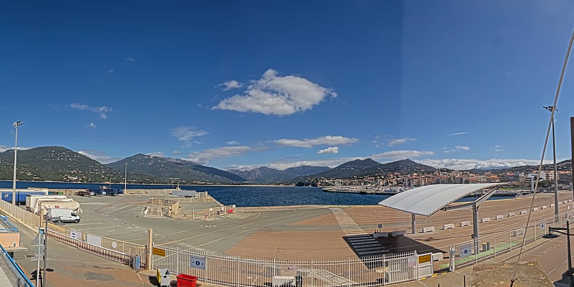 A harbor with a mostly empty parking lot and a covered waiting area, under a mostly sunny sky with a few clouds, is backed by mountains and a town across the water.