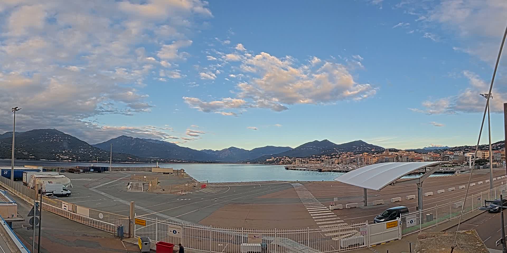 A partly cloudy sky overlooks a harbor with a town and mountains in the background.