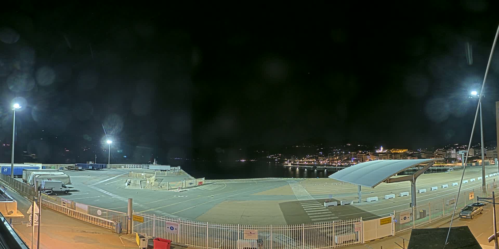 A nighttime view of a mostly empty port area with a few trucks and cars, under a dark, possibly slightly hazy sky, overlooking a town lit up across the water.