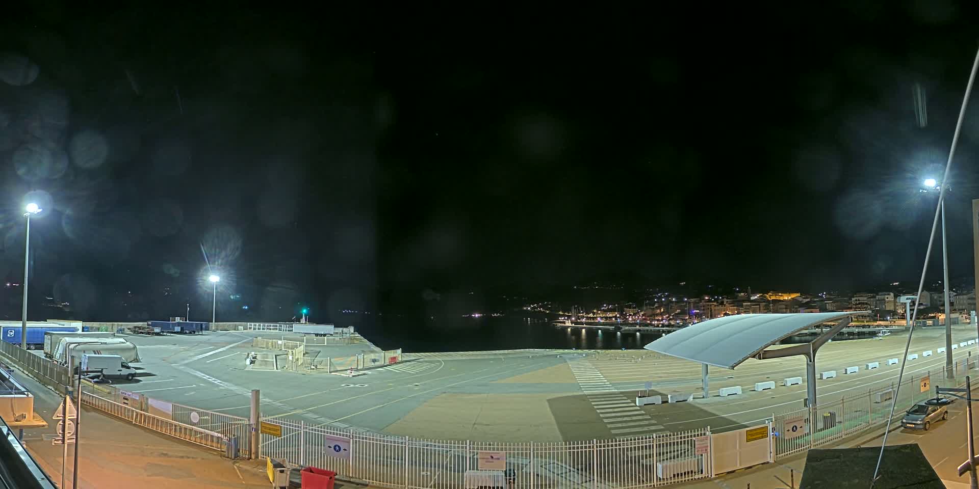 A nighttime view of a mostly empty port area with several parked trucks and cars, under a dark, slightly hazy sky.
