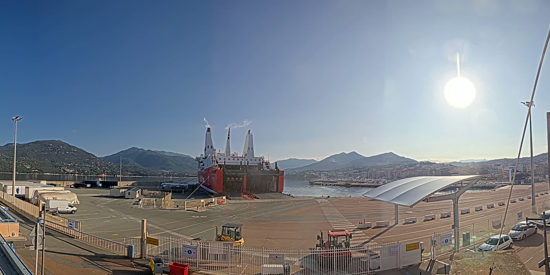 A large red and white cargo ship is docked at a port on a sunny day, with mountains visible in the background.