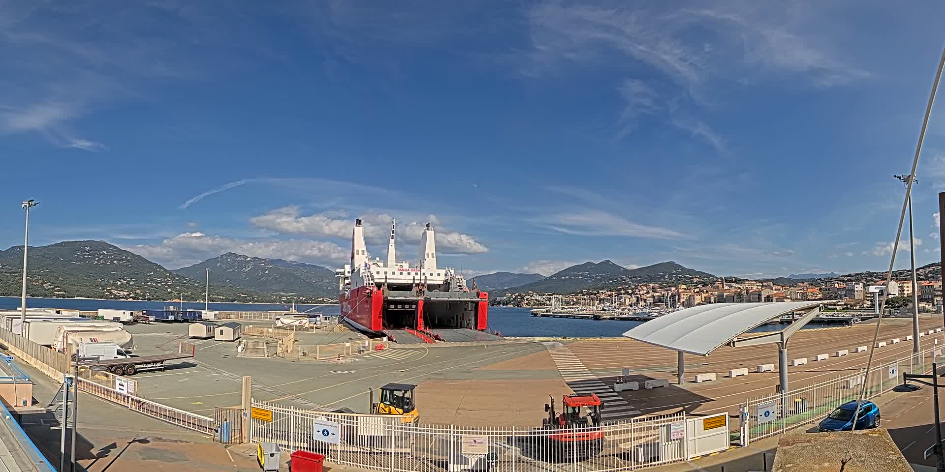 A large red cargo ship is docked at a port under a mostly sunny sky, with mountains in the background.