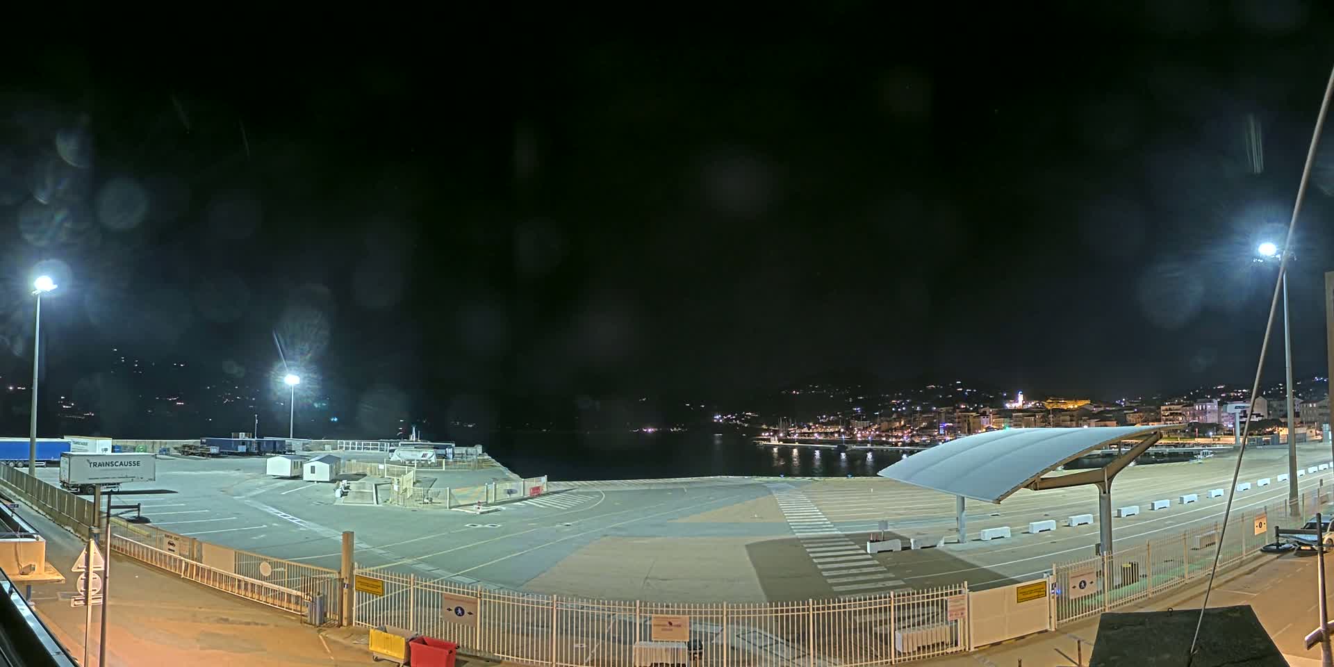 A nighttime view of a mostly empty port area with several semi-trucks and a distant city skyline visible across the water, under a dark, slightly hazy sky.