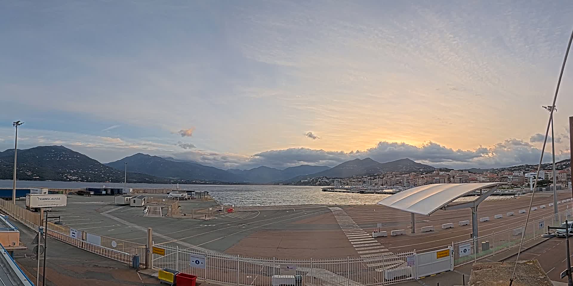 A mostly empty port area with a partially cloudy sunset over mountains and a calm body of water.