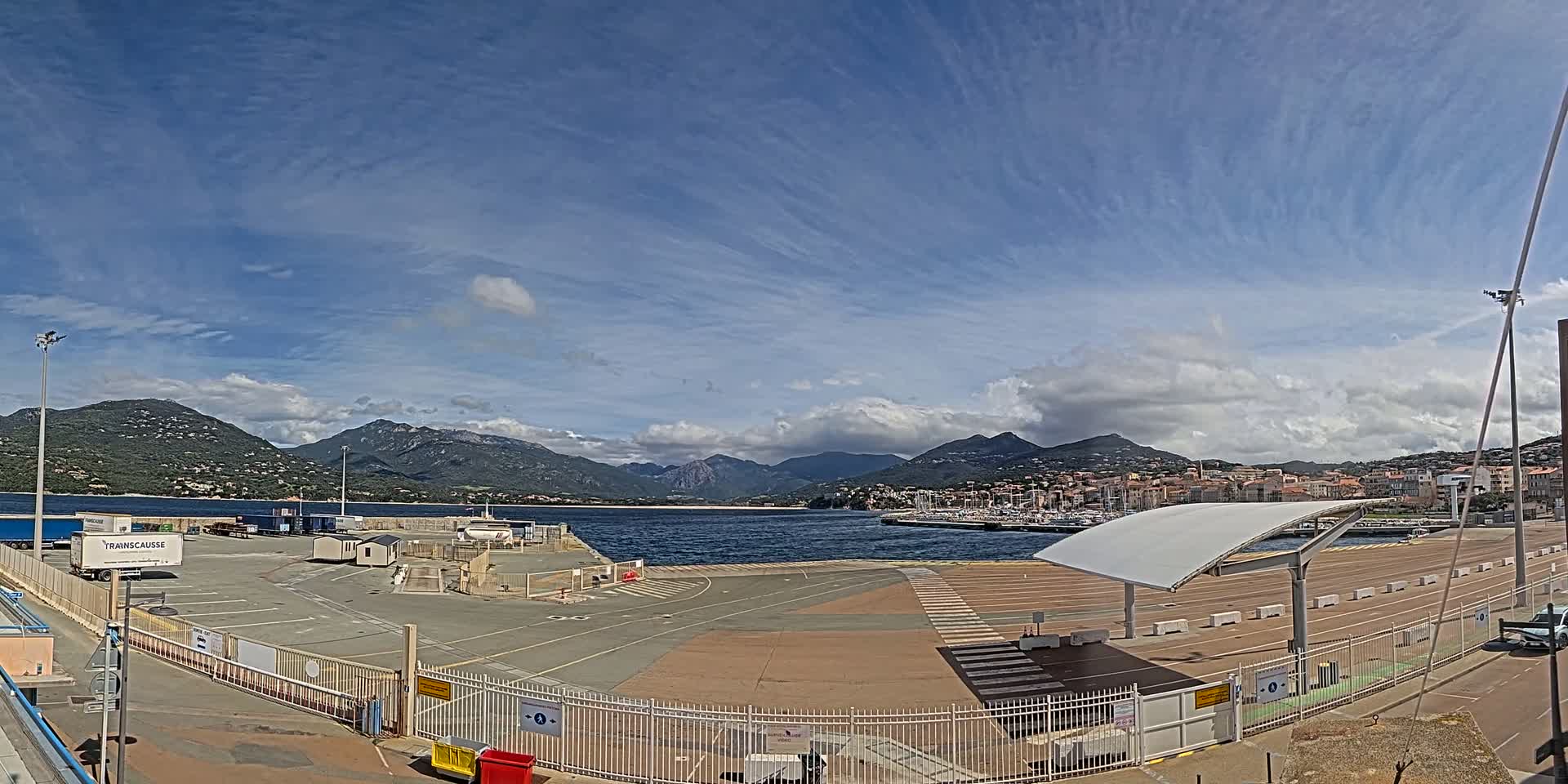 A mostly empty port area with a few parked vehicles, under a partly cloudy sky, overlooks a calm body of water with mountains in the background.