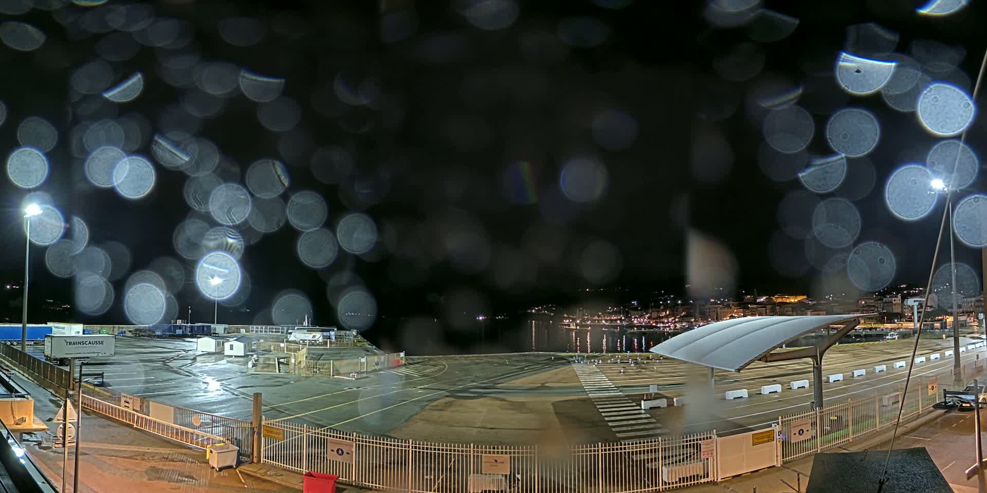 A nighttime view of a port area, seen through rain-covered glass, shows an empty parking lot, a waterfront with docked boats, and city lights in the distance.