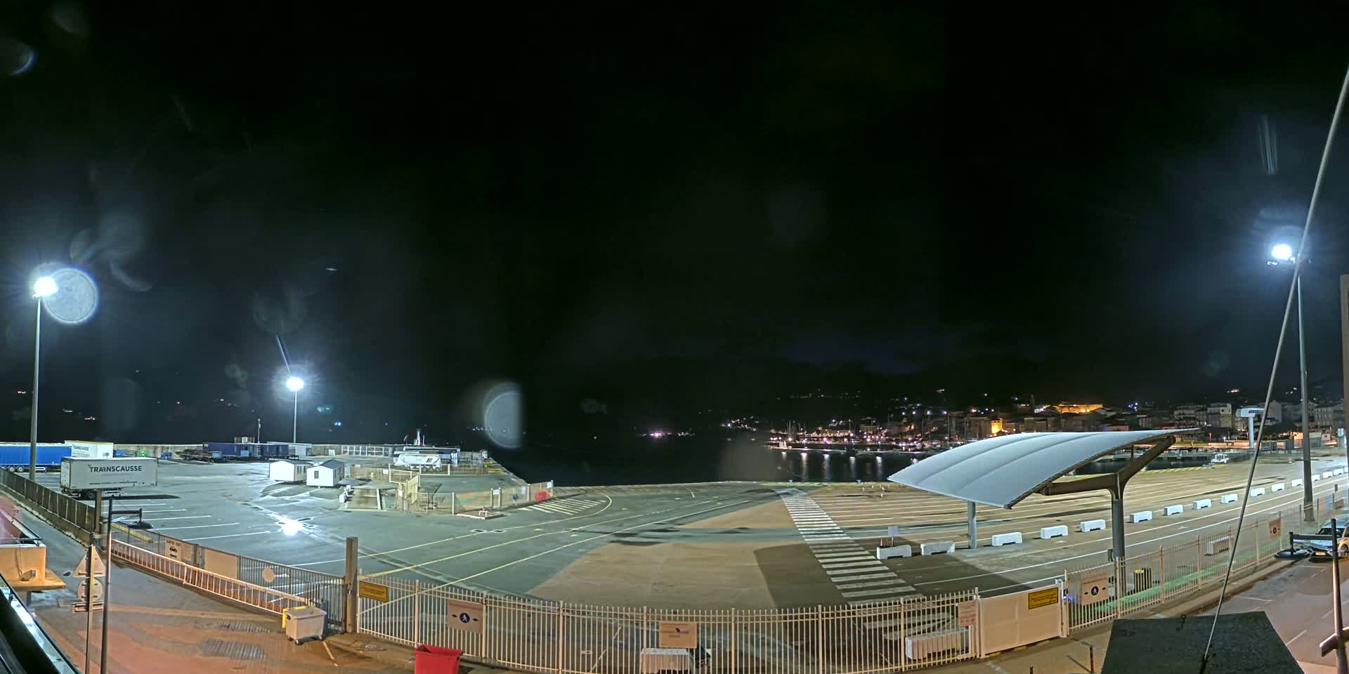 A nighttime view of an empty port area with a few parked trucks and a distant town lit up across the water under a dark, clear sky.