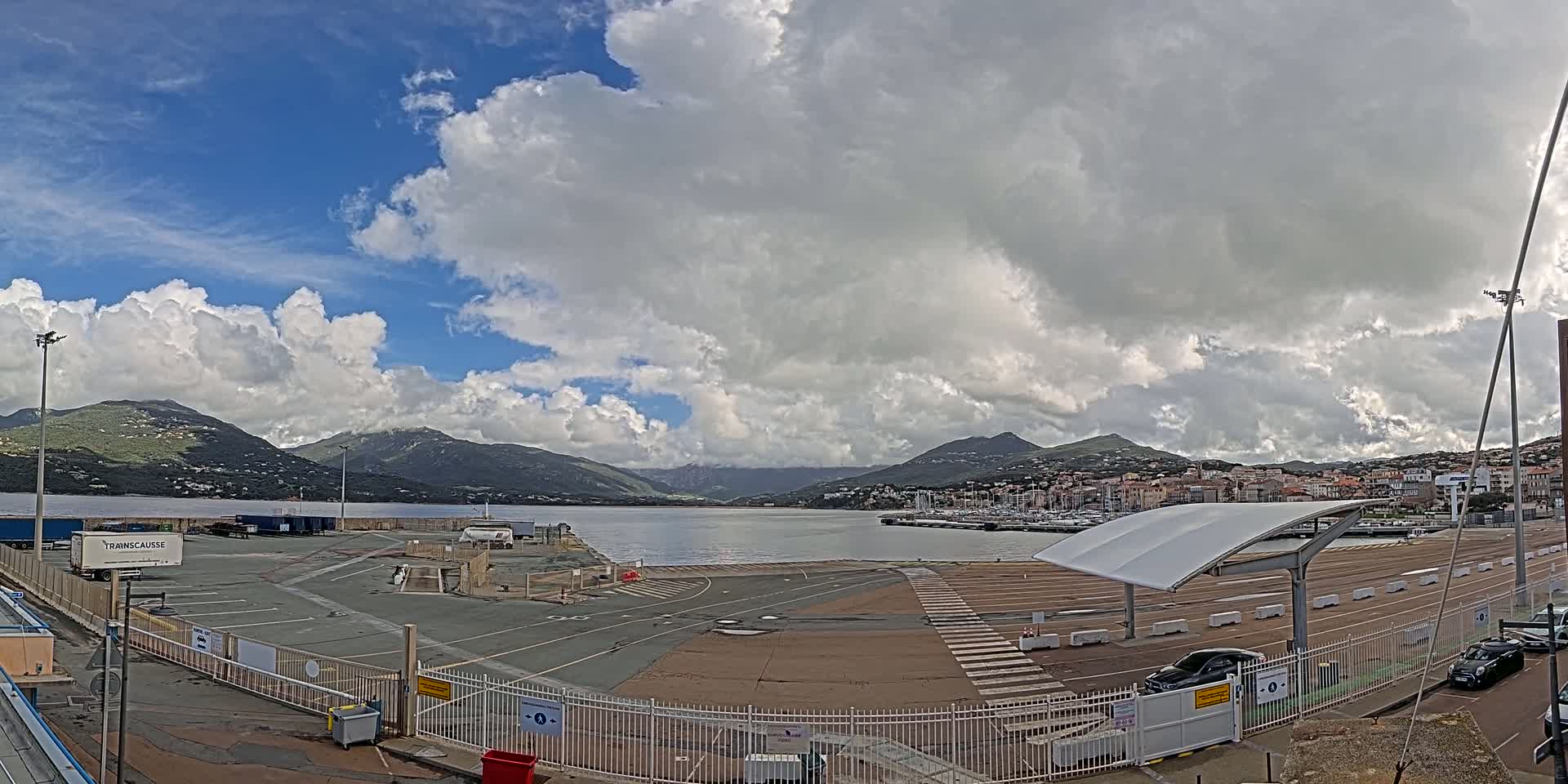 A partly cloudy sky overlooks a port area with a calm body of water, mountains in the distance, and several parked cars.