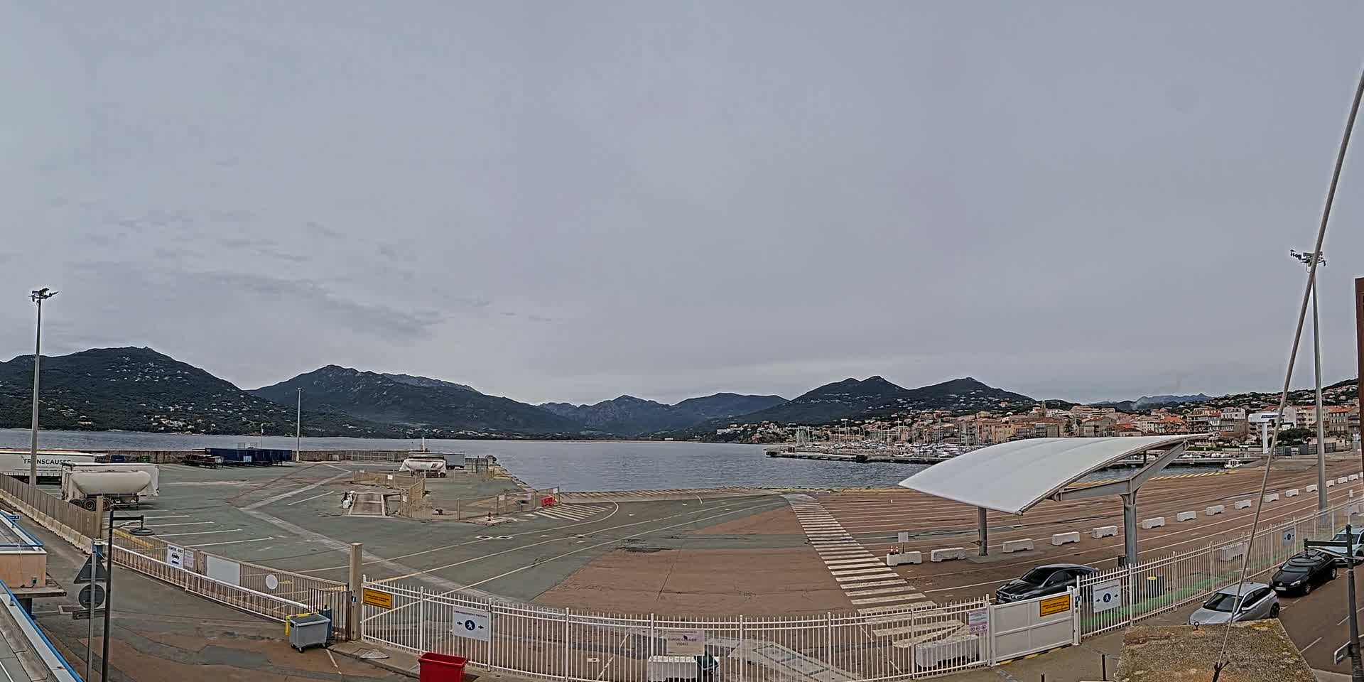 A mostly empty paved port area with a few cars, overlooked by mountains under a cloudy sky.