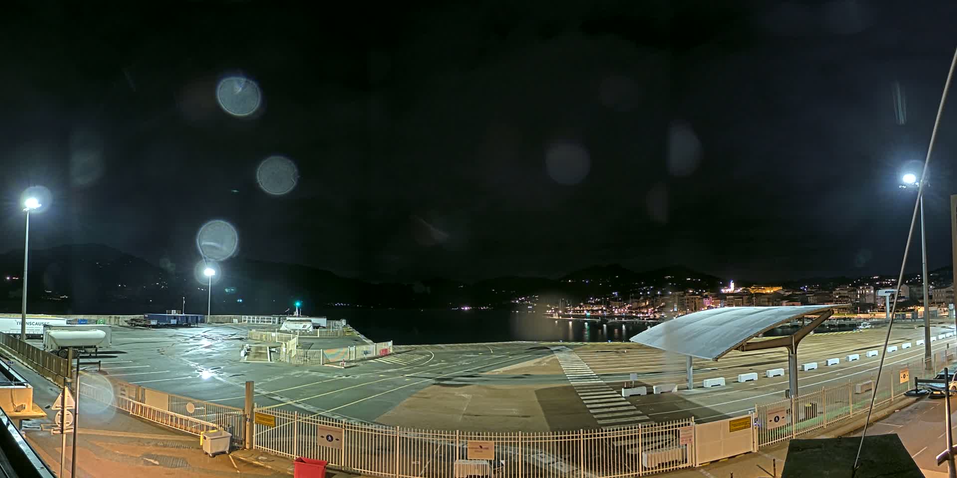 A rain-slicked nighttime port area with a large empty paved lot, a few vehicles, and a distant town illuminated on a hillside across the water.