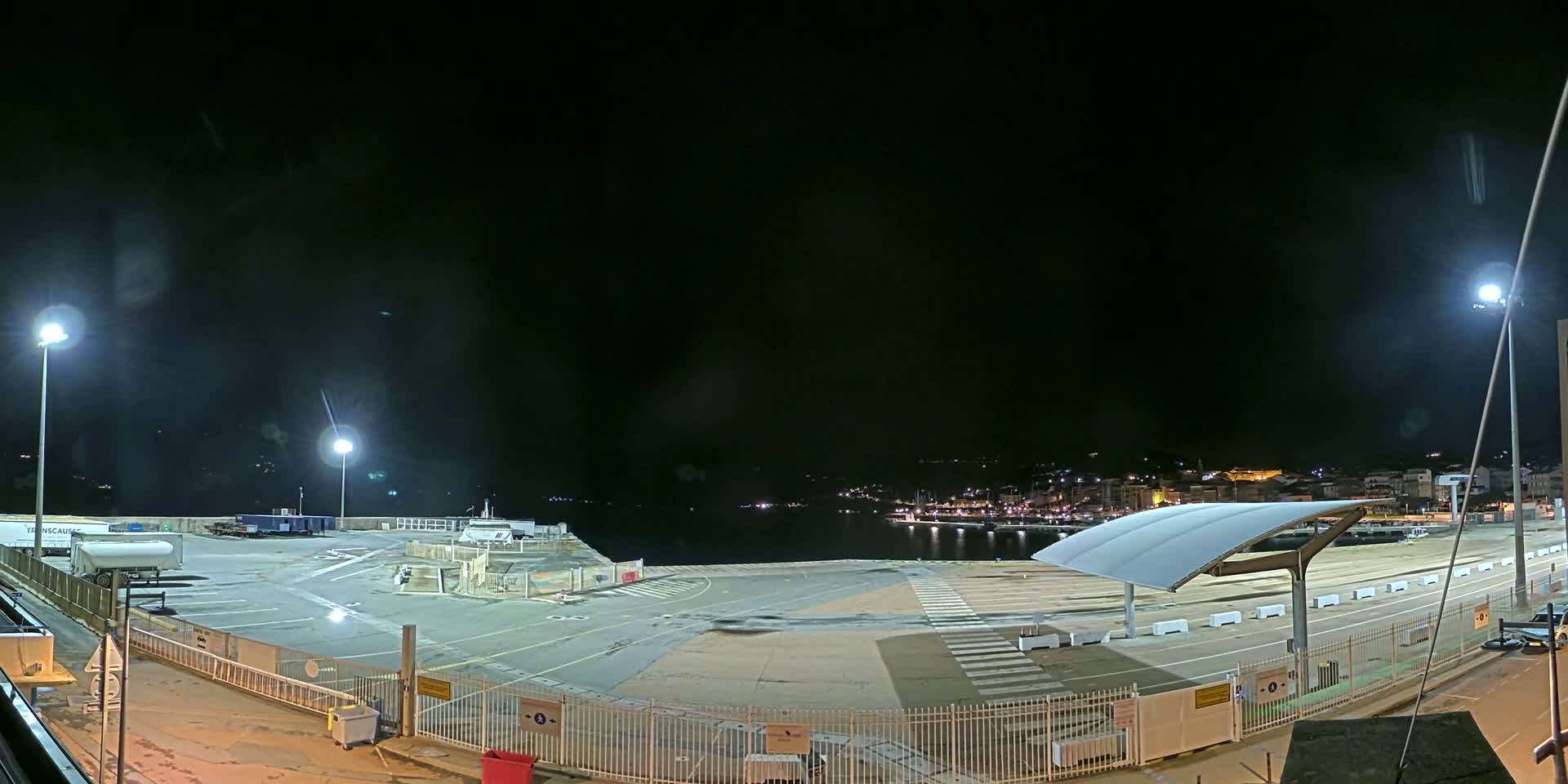 A nighttime view of an empty port area with a few ships and buildings in the distance, under a dark, possibly cloudy sky.