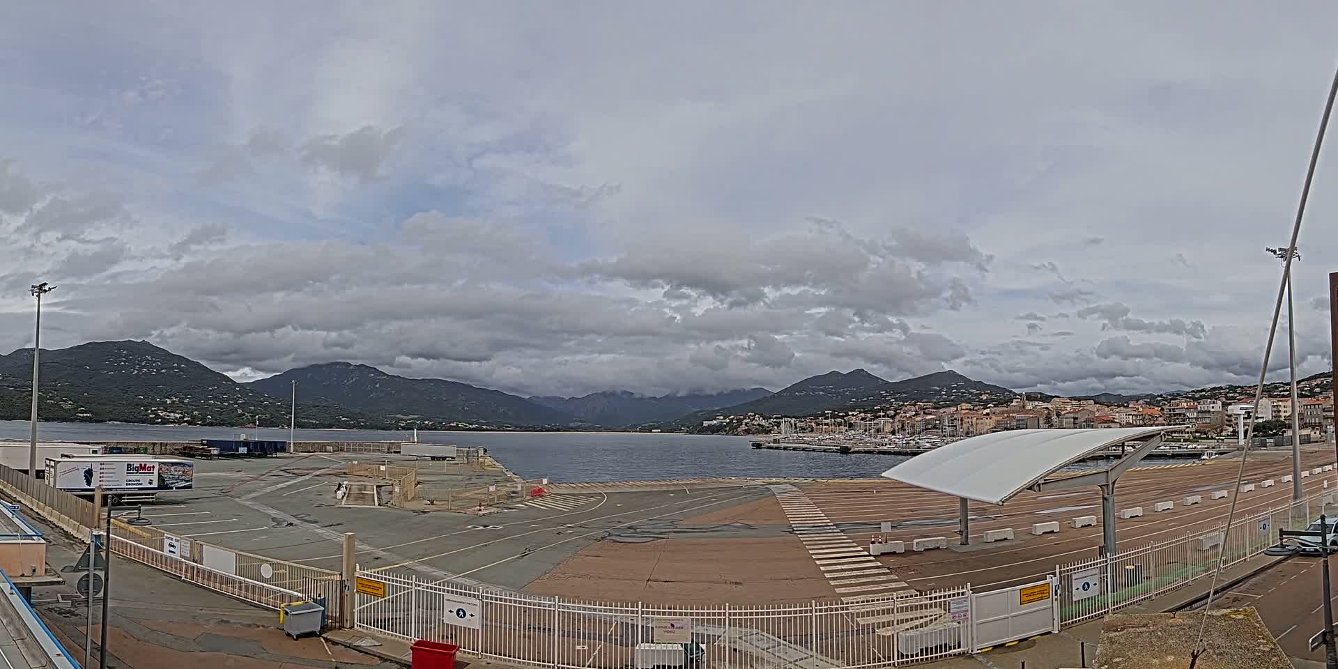 A mostly empty port area with a few vehicles and a white canopy, under a cloudy sky, overlooks a calm body of water and mountains in the distance.
