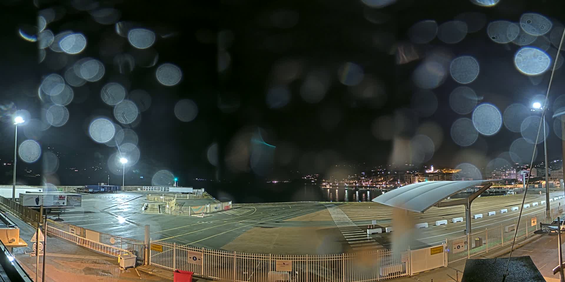 A nighttime view of a rain-soaked harbor area, showing an empty lot, a covered walkway, and a distant cityscape across the water.