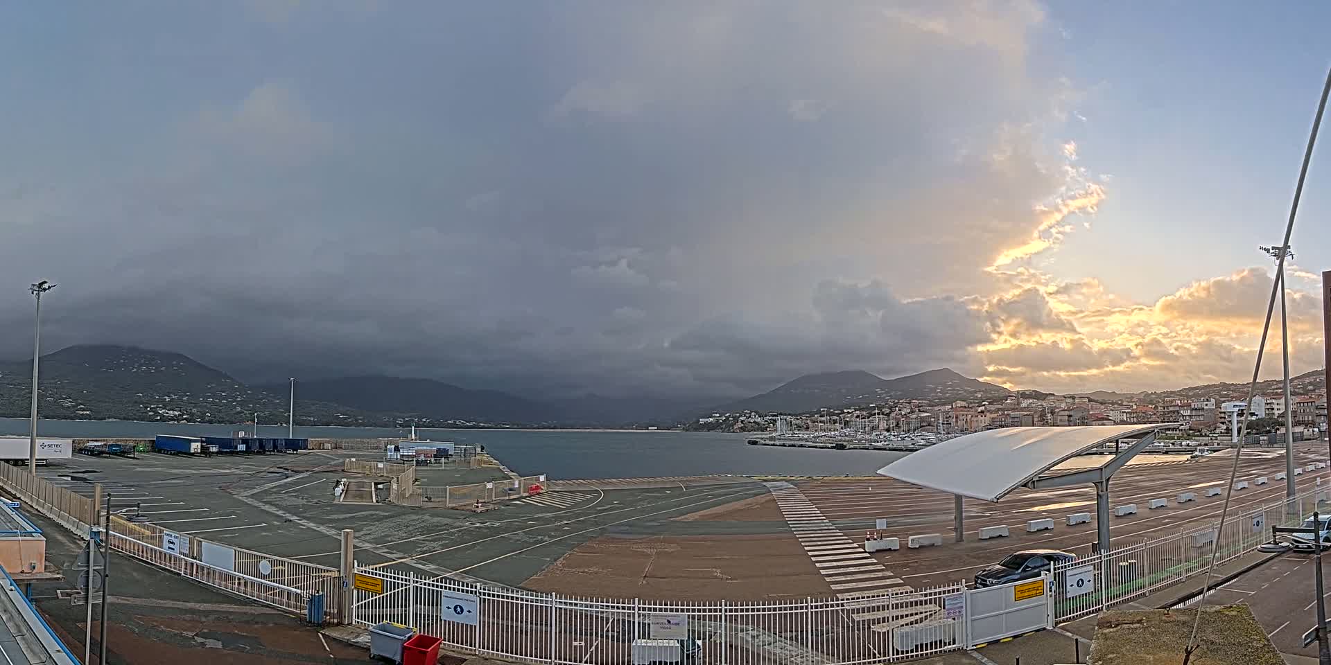 An empty port area with a few cars, under a partly cloudy sky with some golden light breaking through the clouds, overlooks a calm body of water and mountains in the distance.